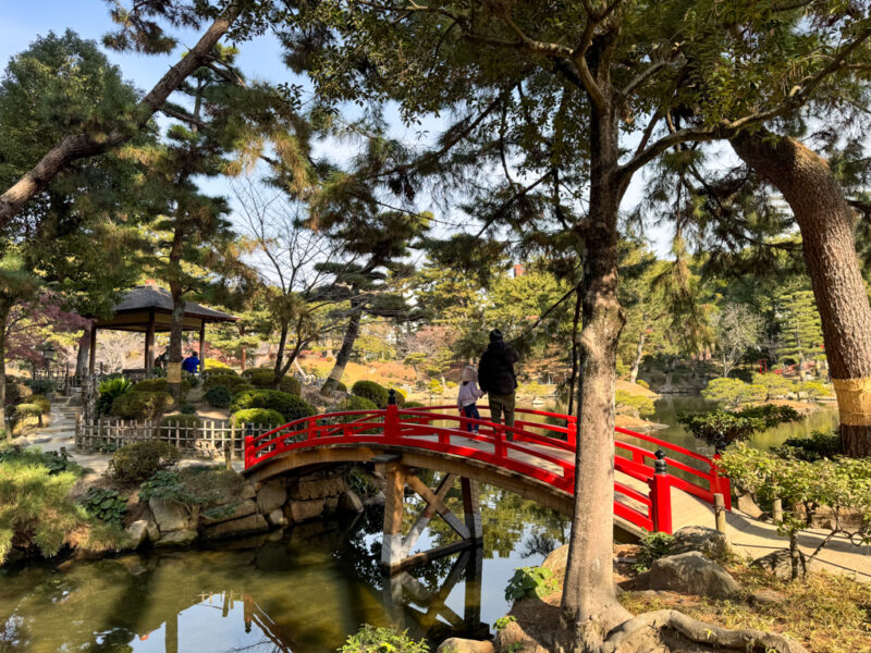 a red bridge over water with people walking on it at Shukkeien Garden in Hiroshima